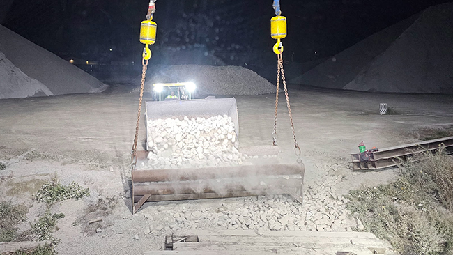 A front-end loader moves cobble on shore at night. The lights of the loader are turned on and facing the camera. The loader is dumping cobble into a swing bucket used to load the cobble onto a barge.